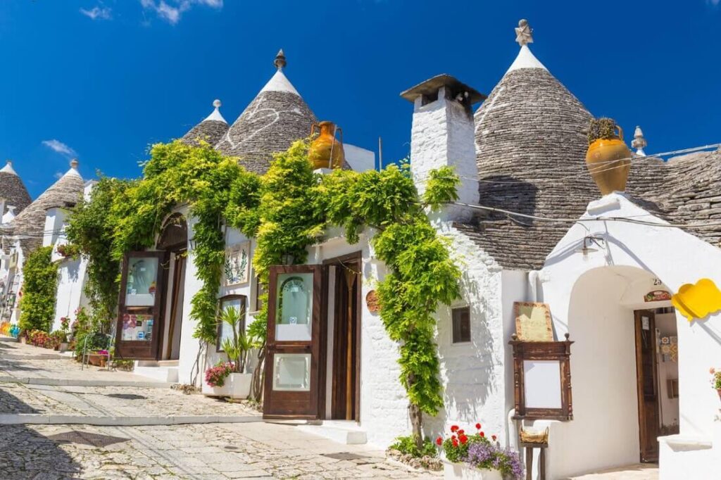 Weiße Trulli mit kegelförmigen Steindächern und grünen Kletterpflanzen an einer gepflasterten Gasse in Alberobello